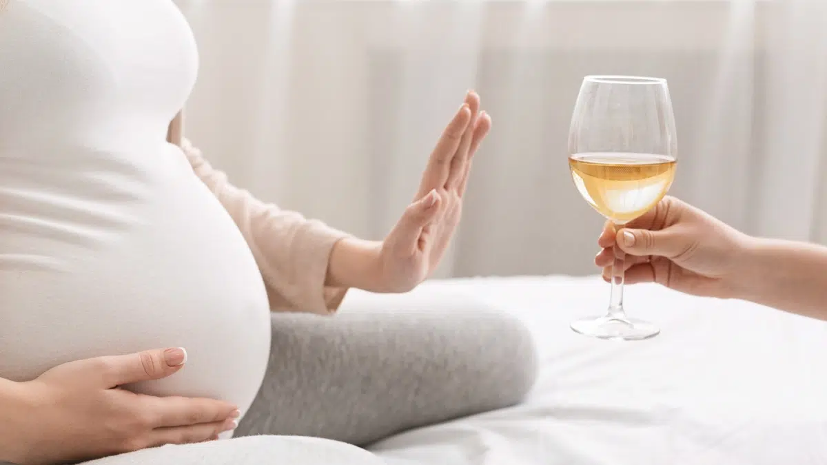 Pregnant woman holding her hand out to reject a glass of white wine. Avoiding alcohol is the safest approach for maternal and fetal health.